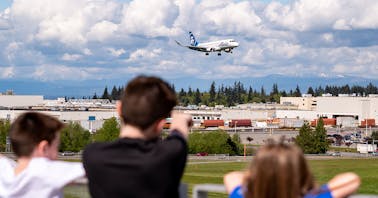 Kids observe a plane landing in Paine Field, with the Boeing Everett Factory and North Cascade mountains in the background.